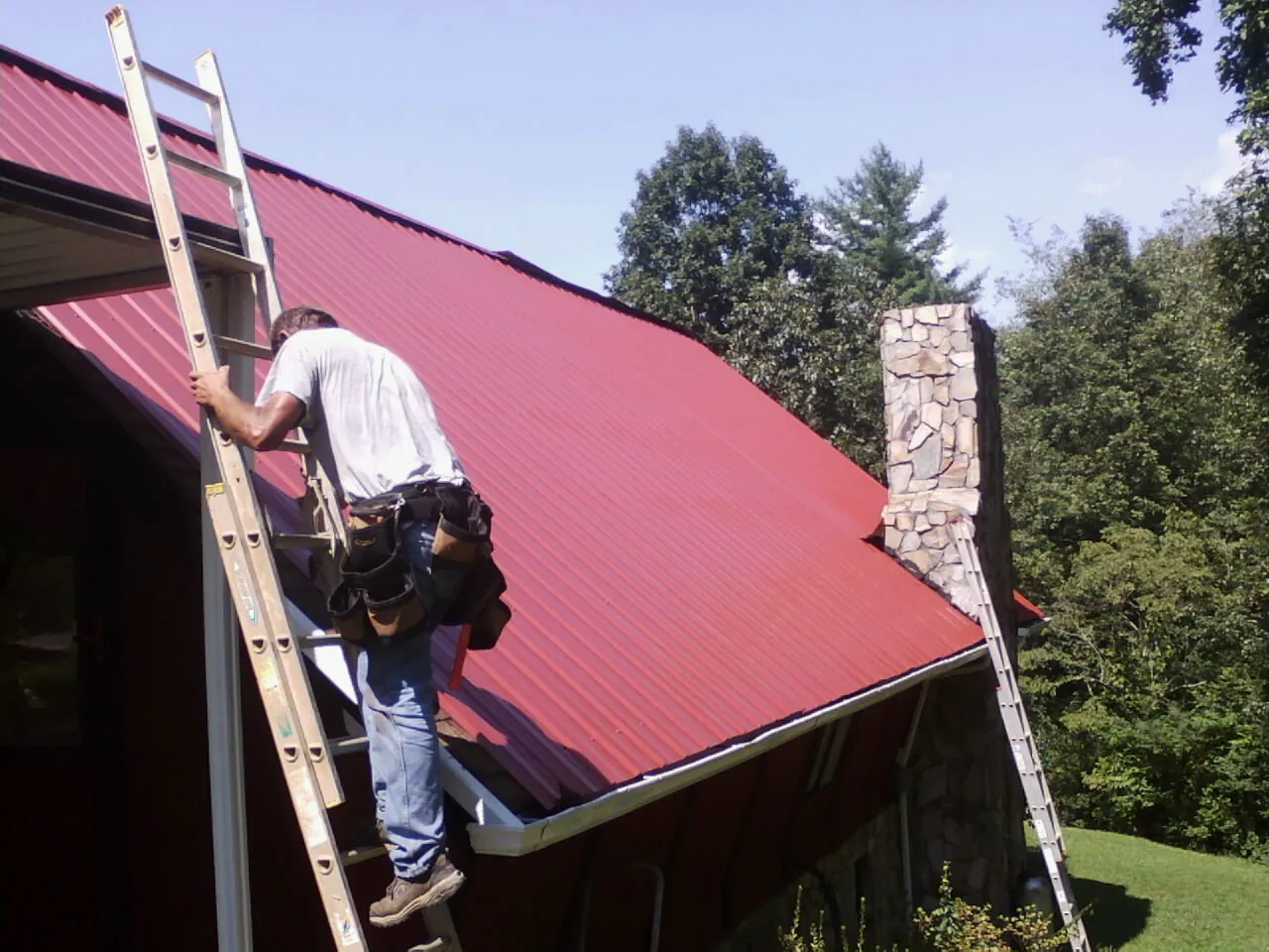 Roofer on a ladder beside a red metal roof and stone chimney — Soffit & Fascia Repair by Roofing Brothers in Peninsula
